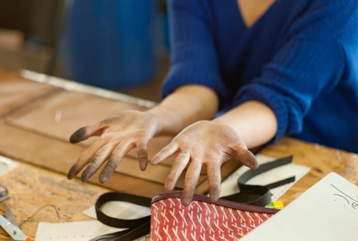 Close-up of a maker's ink-stained hands working with leather pieces, a zipper pouch, and strap components on a wooden workbench.