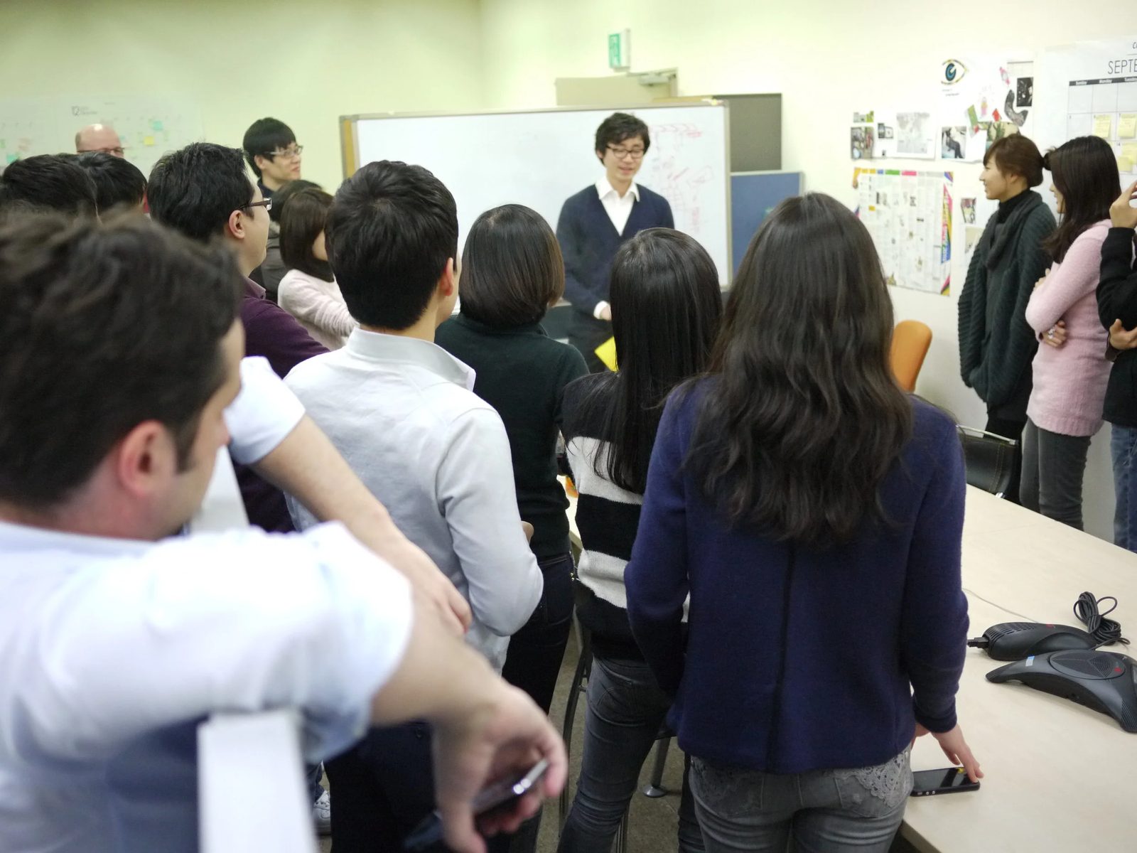 Innovation workshop in session: a large group of participants gathered around a facilitator at a whiteboard, with idea maps and notes pinned to the walls behind them.